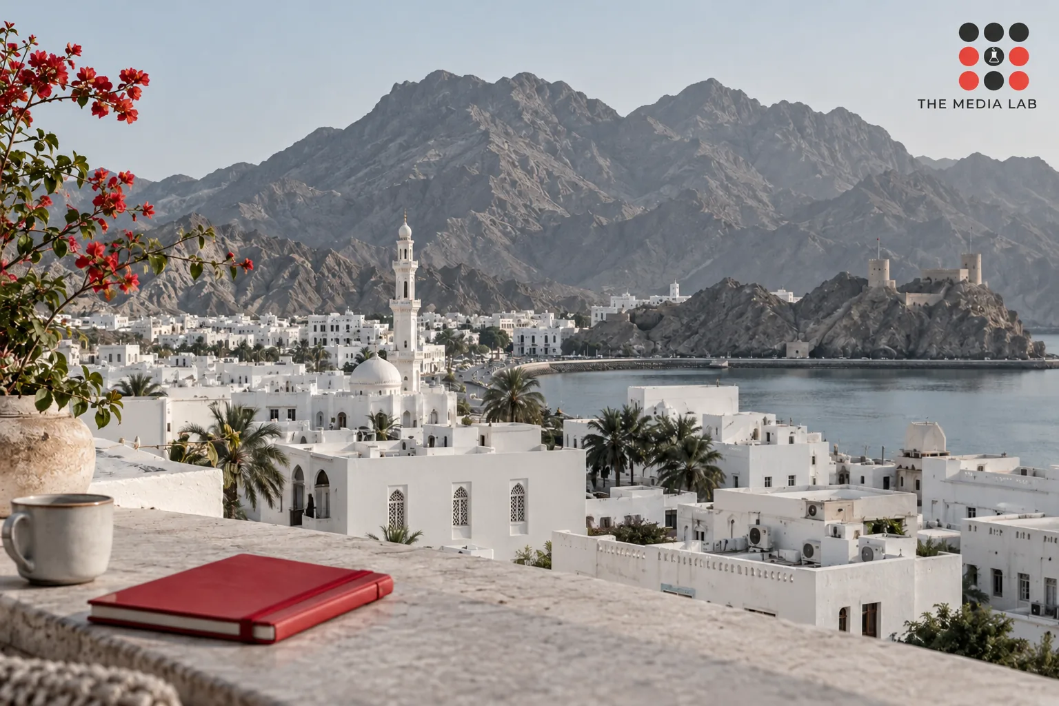 Architectural editorial photograph of Muscat white buildings and rugged grey mountains, subtle red notebook in foreground, no Dubai skyline — The Media Lab