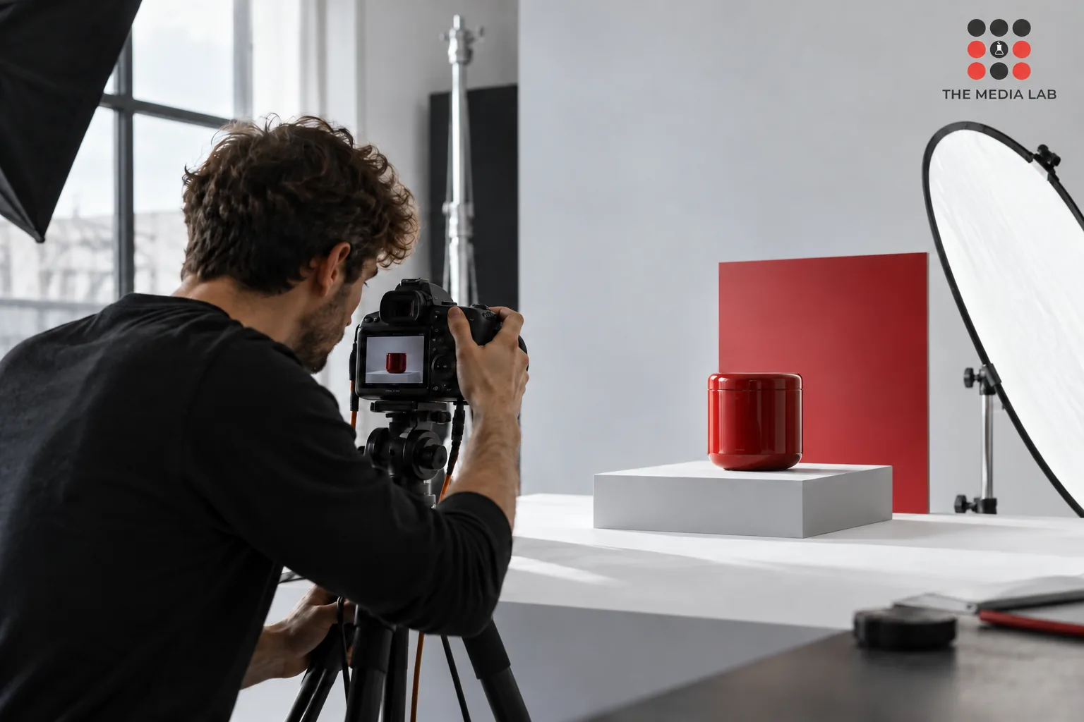 Photographer adjusting a studio camera beside a red product and white reflector — The Media Lab